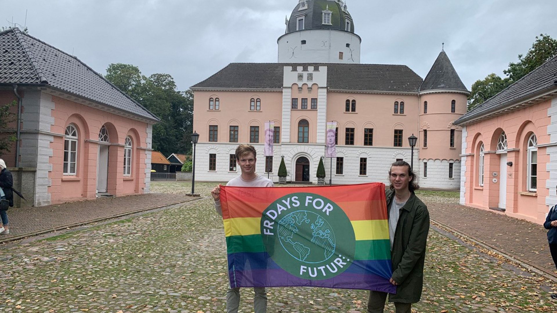 Auf dem Bild stehen Jonas und Janno vor dem Schloss in Jever, sie halten eine Flagge mit dem FFF-Logo vor einer Pride-Flagge